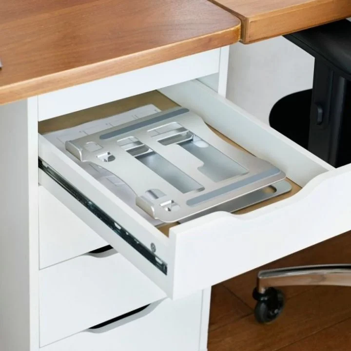 白い引き出しが開いており、その中に銀色の折りたたみ式ノートパソコンスタンドが収納されている様子。木製の天板と白い収納家具の一部が見える。
