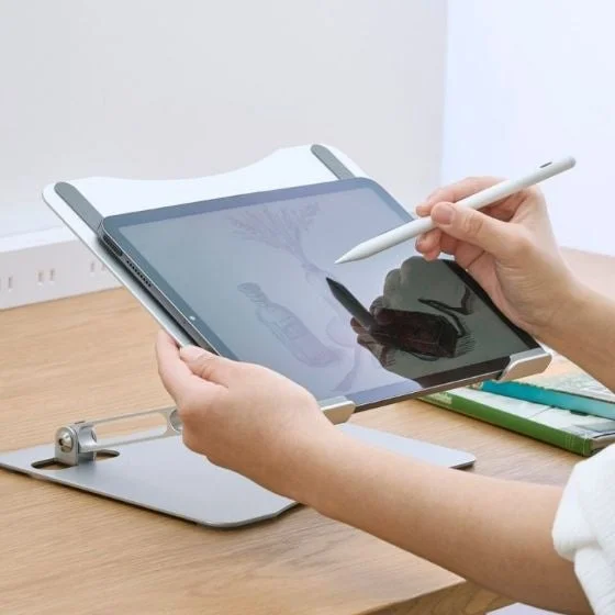 木製のデスクで、調整可能なスタンドに置かれたタブレットをスタイラスペンで操作している人物の手元を捉えた画像です。タブレットの画面にはデジタルアートのようなものが表示されており、集中して作業している様子がうかがえます。