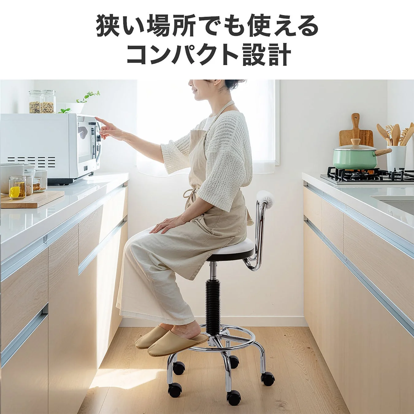 狭いキッチンで女性がコンパクトなキャスター付きスツールに座り、電子レンジを操作している様子