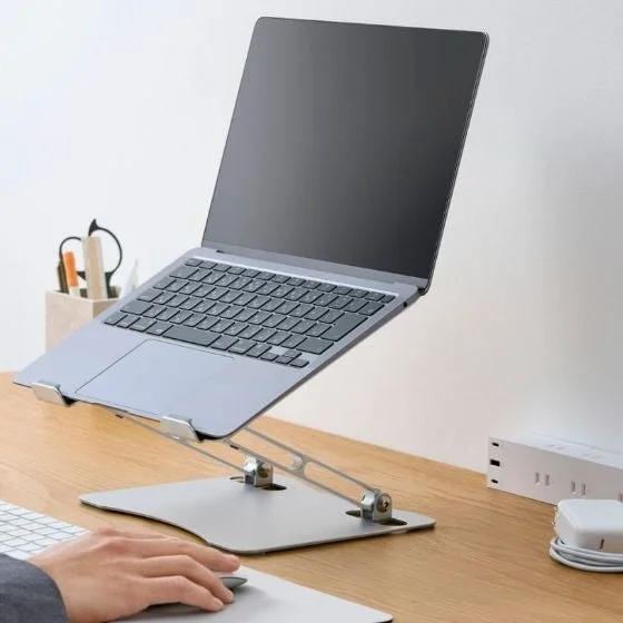 シルバーのノートパソコンが調整可能なラップトップスタンドに置かれ、木製のデスクで使用されている様子。手前にはマウスを操作する手があり、効率的な作業環境がうかがえます。