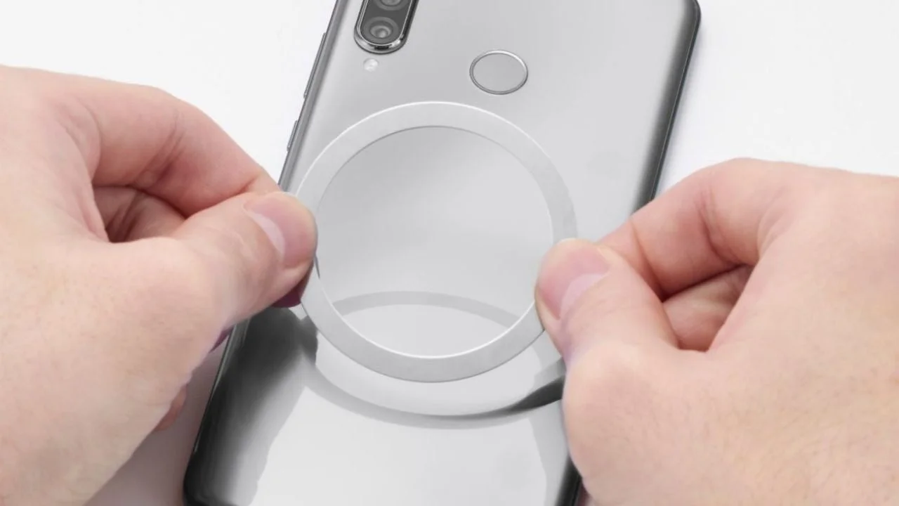 スマートフォンの背面に、円形の白いリング状のアクセサリーを貼り付けようとしている様子です。
